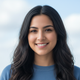 Close-up portrait of a smiling young woman with dark hair, looking at the camera, against a soft blue and white background.